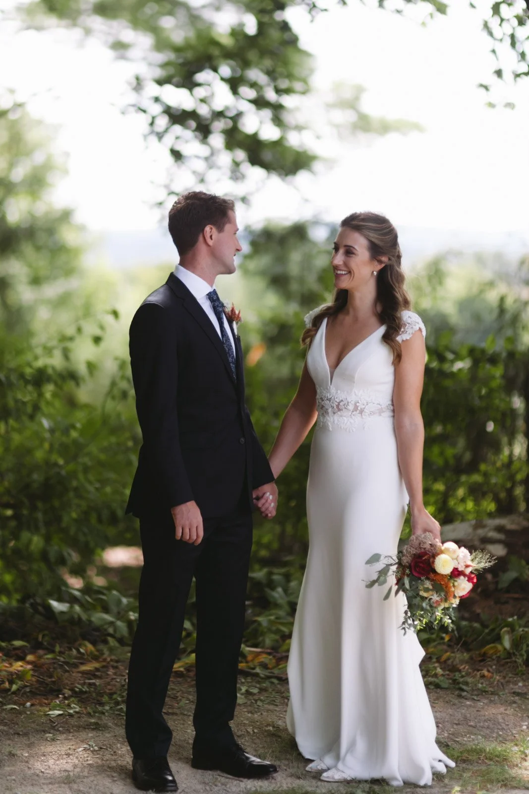 A bride and groom holding hands, standing outdoors in a wooded area, facing each other and smiling. The bride is wearing a white wedding dress with lace details and holding a bouquet of flowers, while the groom is dressed in a dark suit with a tie.