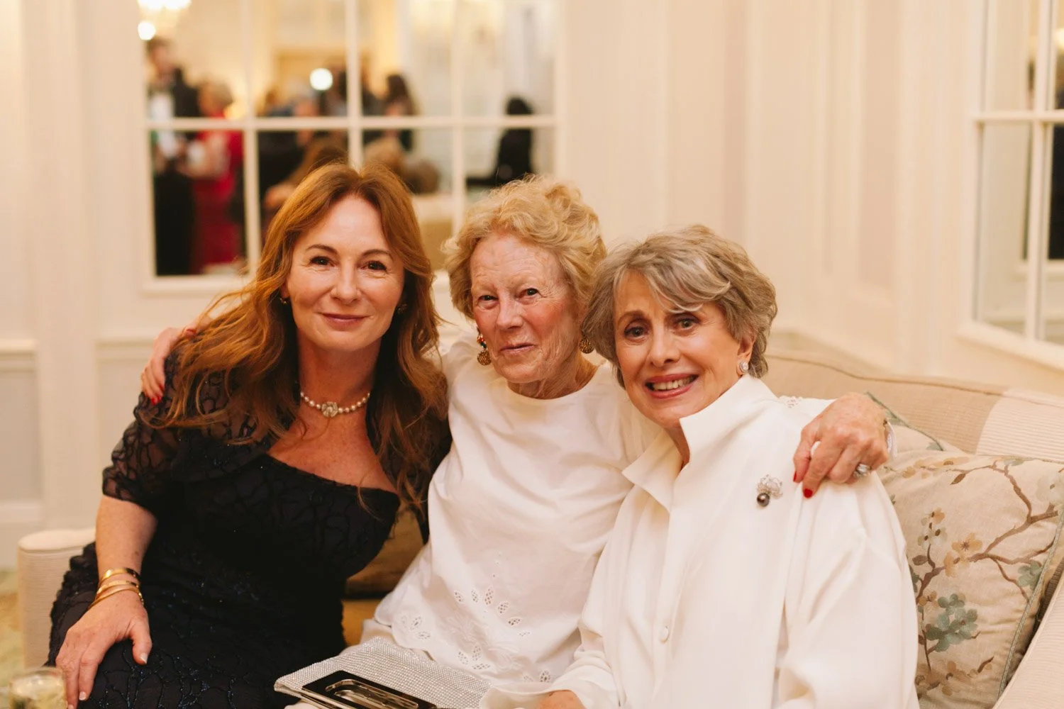 Three women sitting closely on a sofa at an indoor event, smiling and looking at the camera.