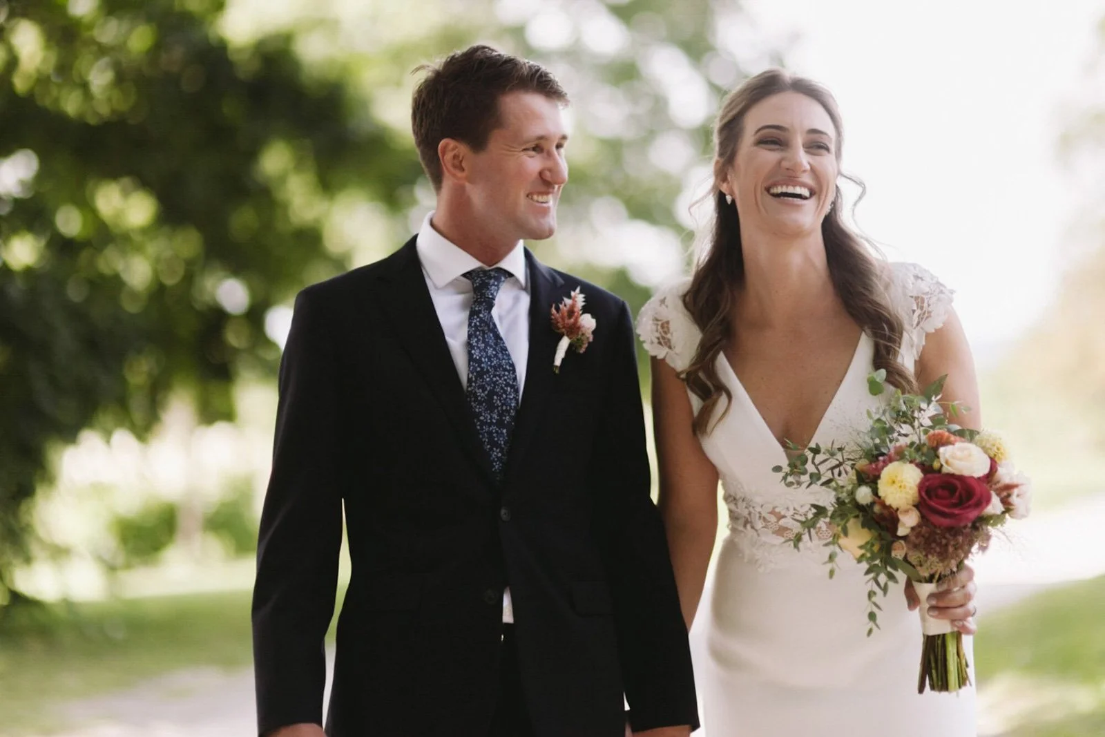 A bride and groom walking outside, smiling and holding hands, with the bride holding a bouquet of colorful flowersat Court Hill in The Berkshires.