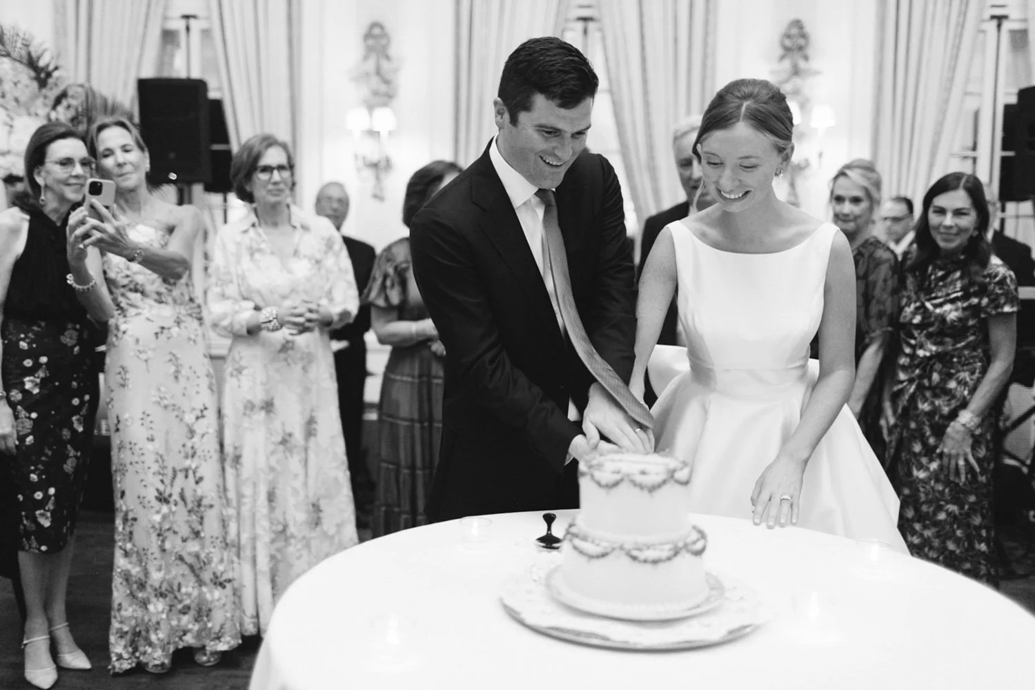 A bride and groom cut a wedding cake together at their wedding reception, surrounded by guests.