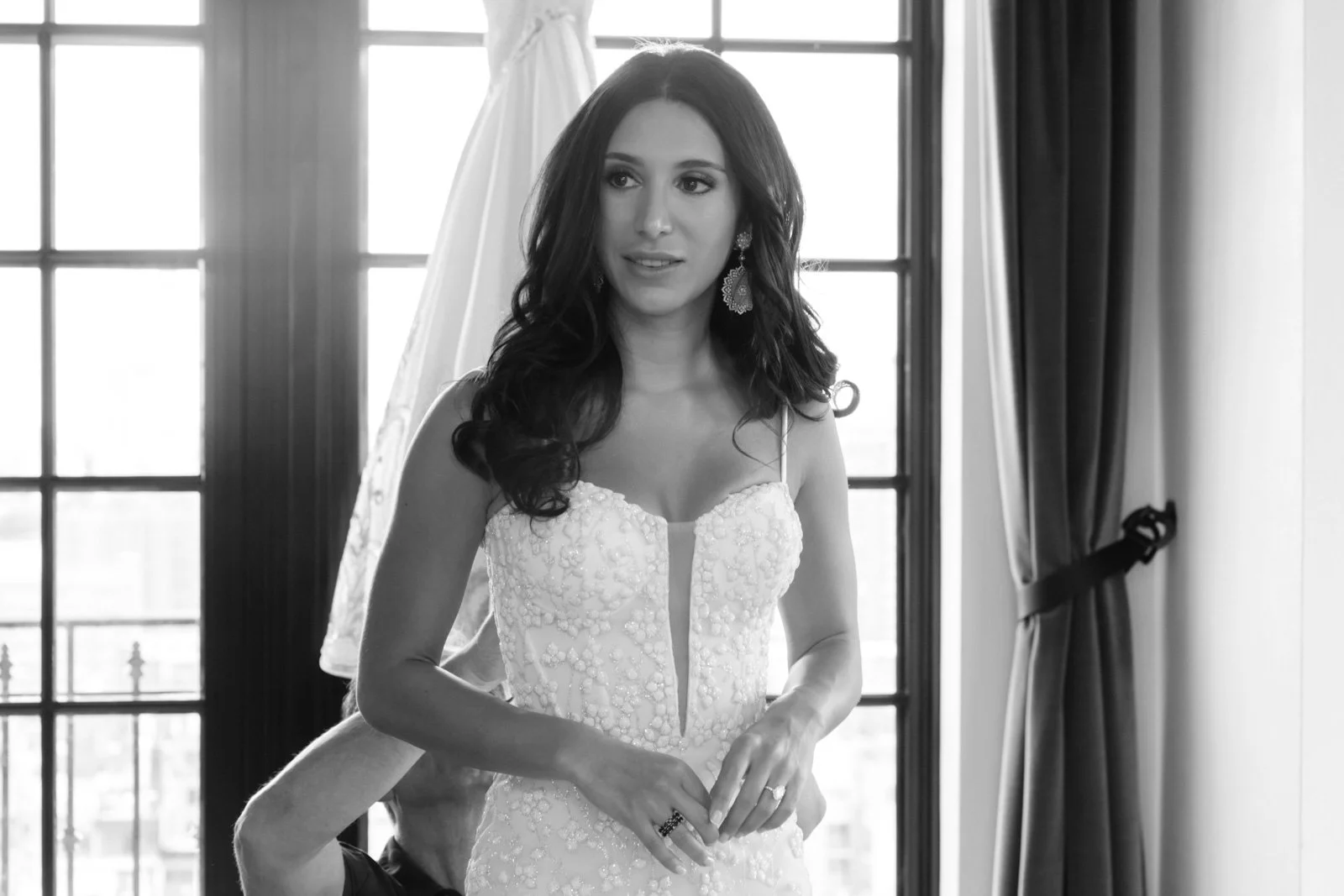 A woman in a wedding dress getting ready, standing in front of large windows with curtains.