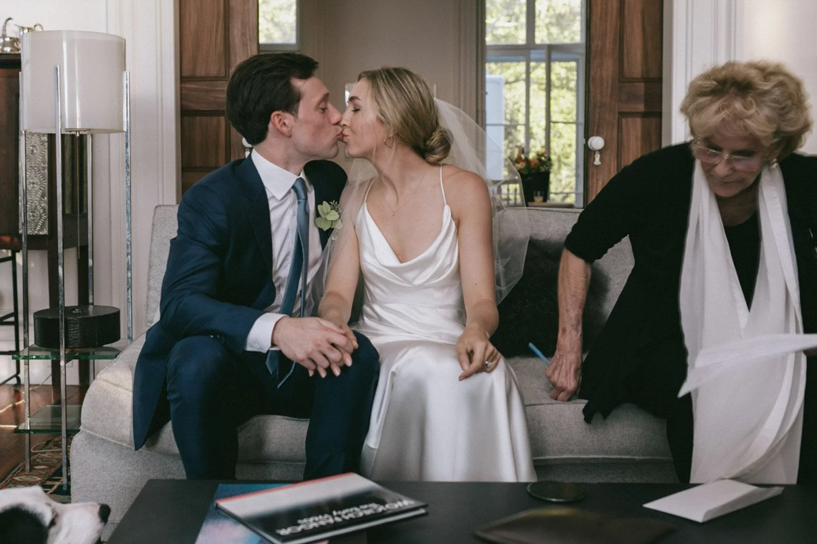 A newlywed couple sharing a kiss on a sofa while sitting indoors, with an older woman leaning over a table nearby.