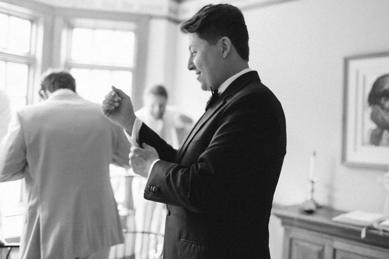 A young man in a tuxedo adjusting his cufflink in a bright room, with other people in the background, including a person in a white suit with their back turned.
