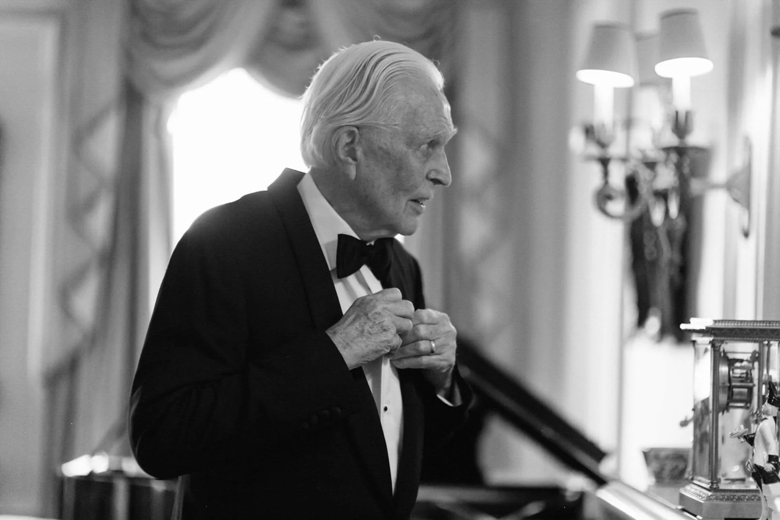 An elderly man in a tuxedo adjusting his bow tie in a room with ornate curtains and a candelabra on the wall.