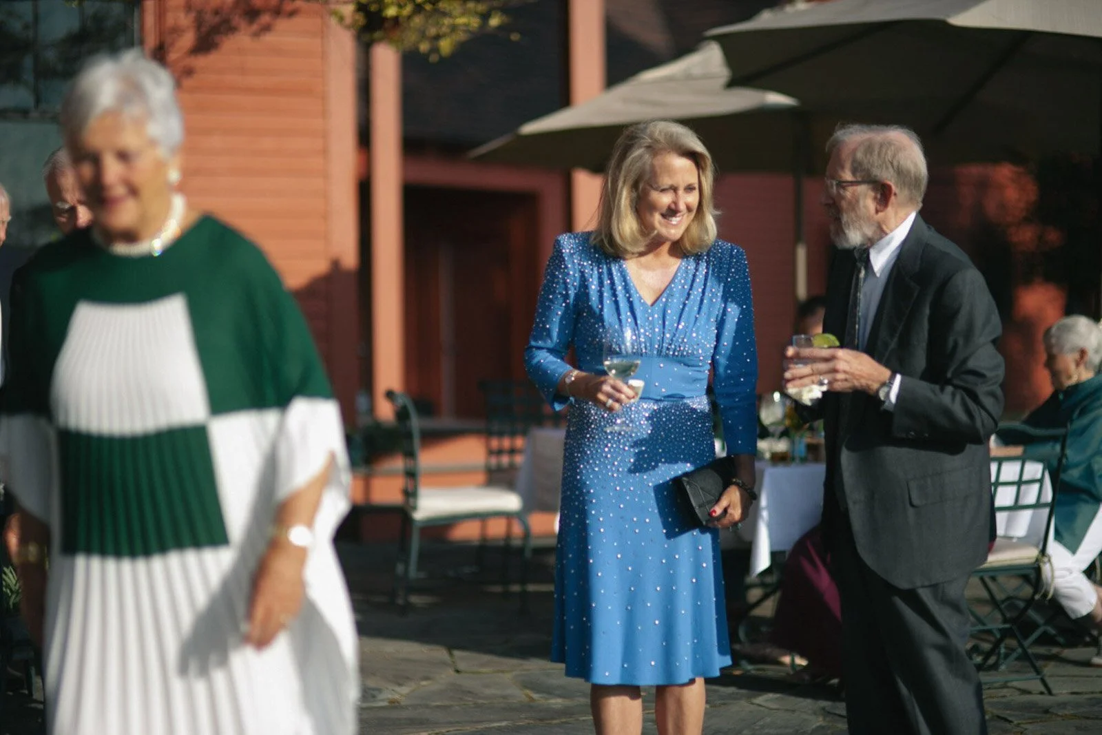 People socializing outdoors at a gathering, wearing formal attire, with a woman in a blue dress and a man in a suit holding drinks, under a patio umbrella.