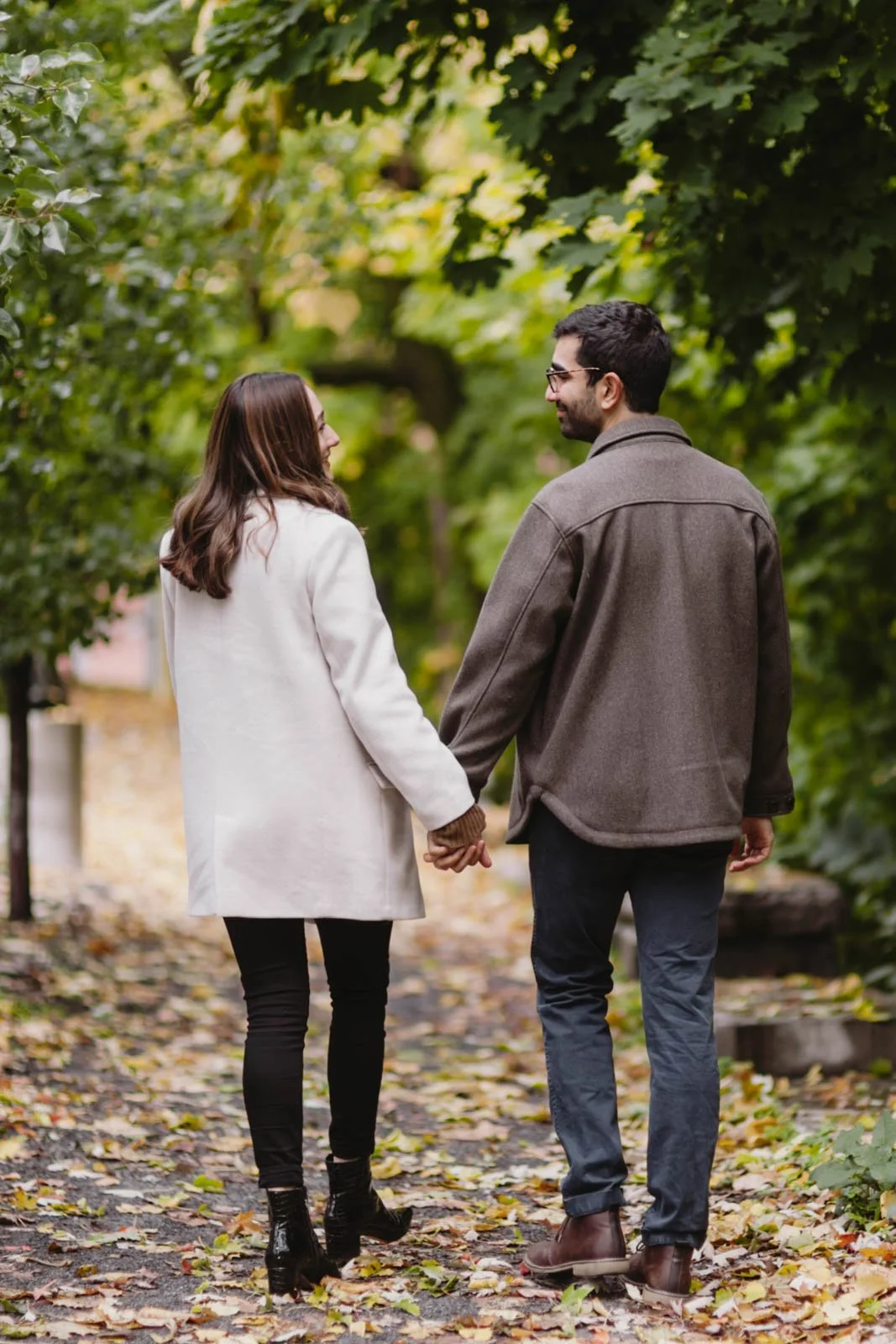 A couple holding hands and walking on a leaf-covered path in a park or forest, surrounded by green trees