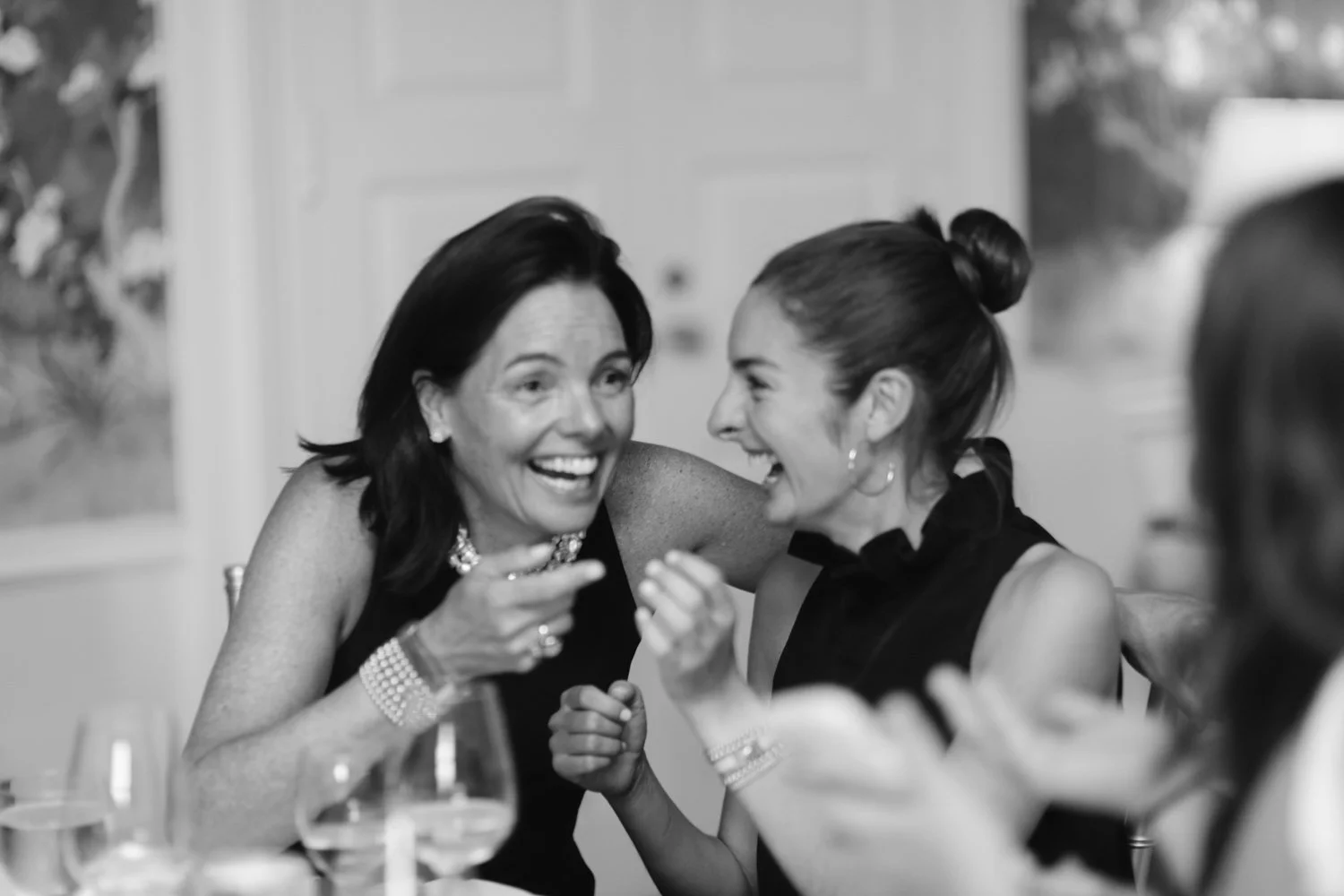 Two women smiling and laughing at a dinner table, engaged in conversation. One is older with dark hair, the other younger with her hair in a bun. Both are dressed up and wearing jewelry.