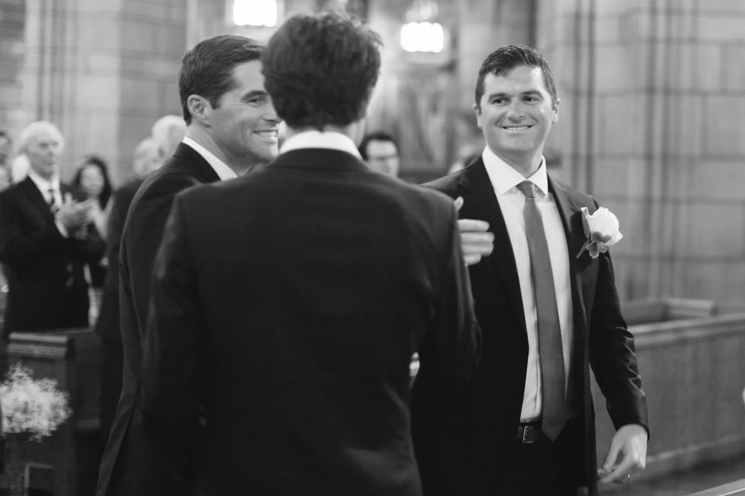 Three men dressed in suits and ties, smiling, at a formal event or ceremony inside a wood-paneled room, with other people in the background.