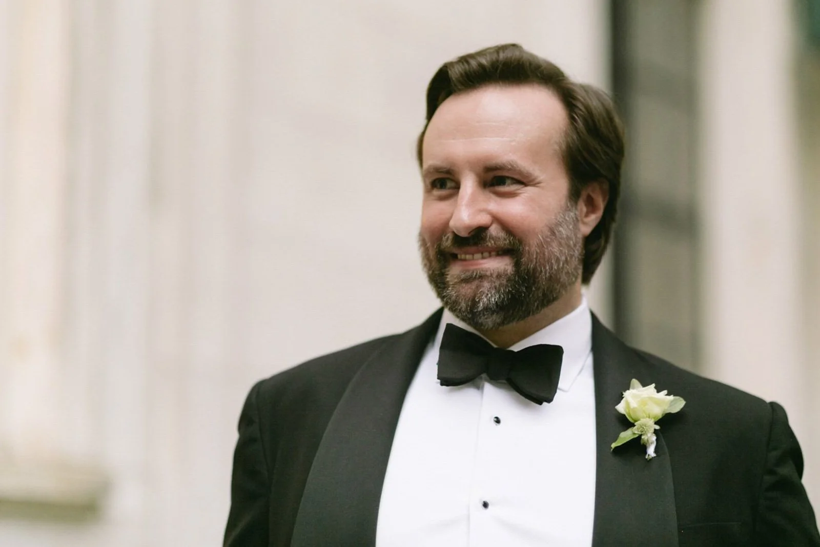 A man wearing a tuxedo with a bow tie and a white boutonniere, smiling and looking to the side indoors.