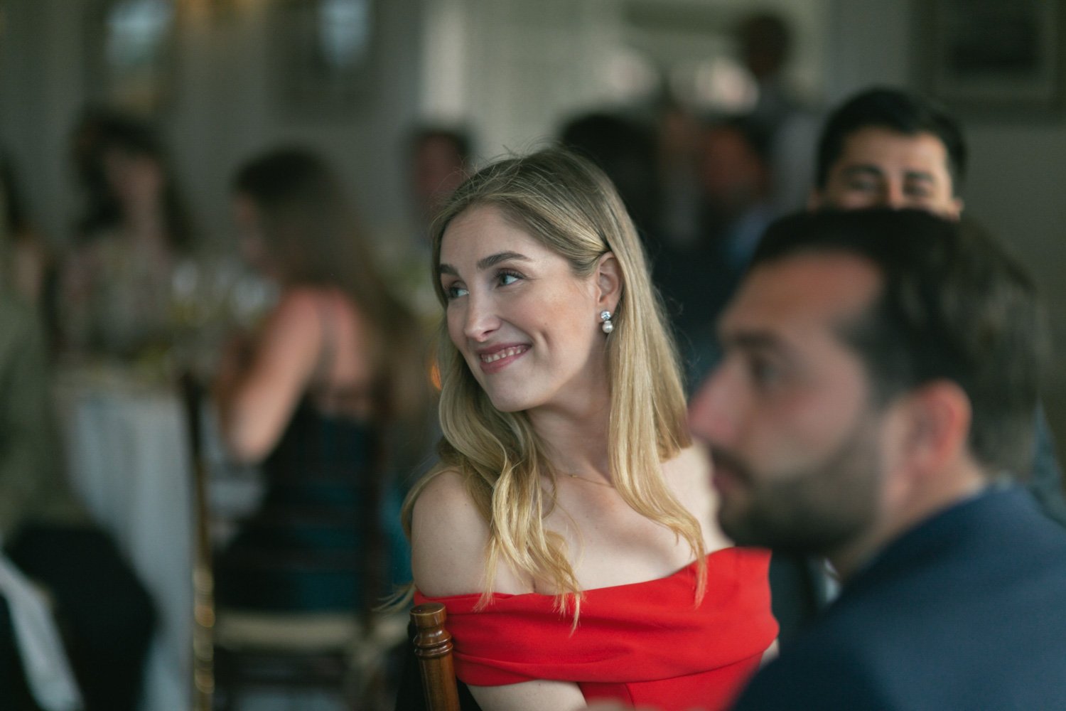 A woman in a red off-the-shoulder dress smiling at a social gathering with other people in the background.