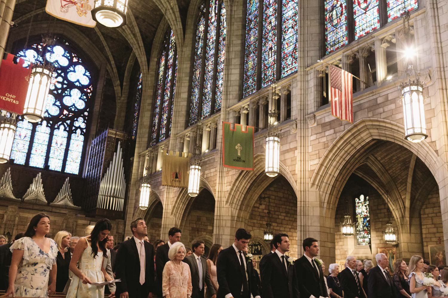 People attending a wedding ceremony inside a large Gothic-style church with stained glass windows and hanging banners.