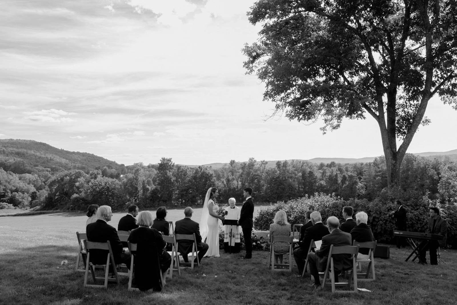 A wedding ceremony outdoors in a grassy field with a large tree and scenic hills in the background. The bride and groom stand facing each other at the altar, surrounded by seated guests, with a musician playing keyboard nearby.
