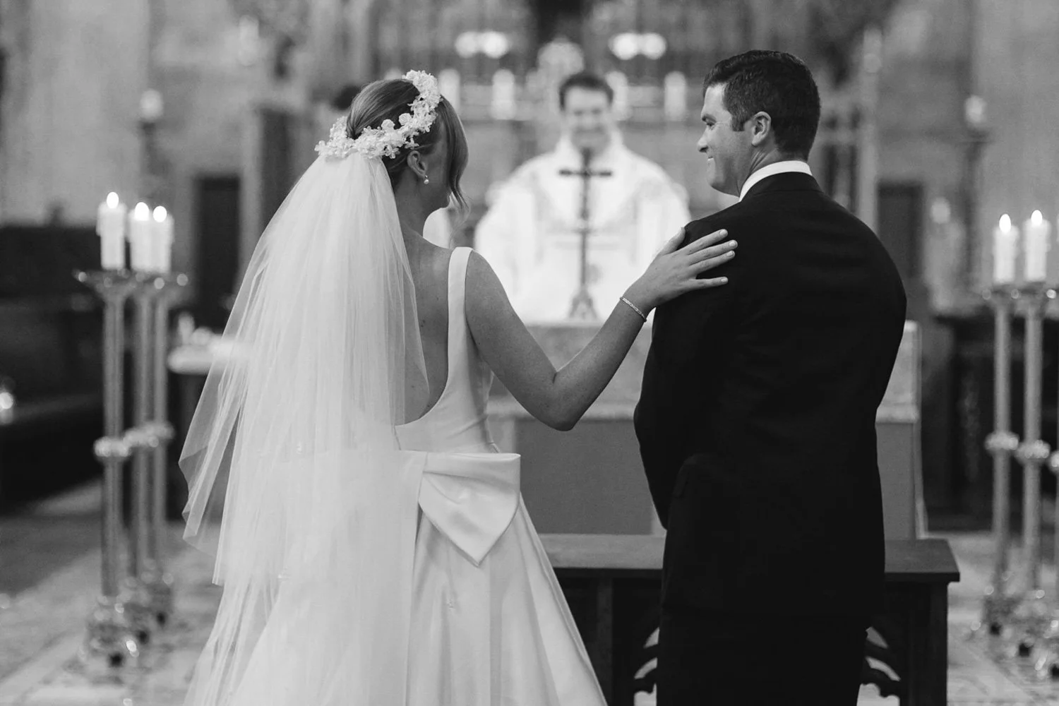 A bride and groom standing at the altar during their wedding ceremony inside a church, with candles and an officiant in the background.
