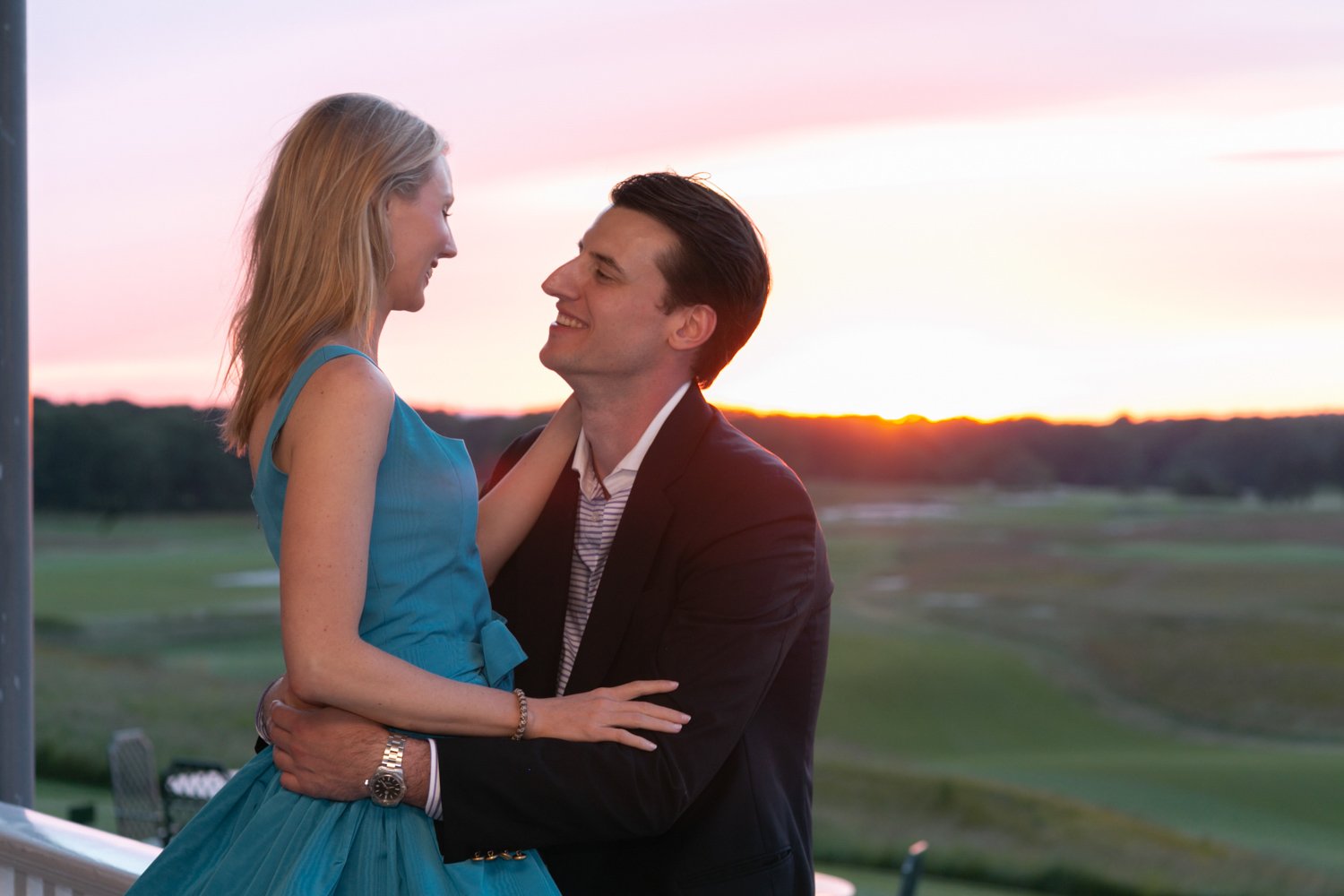 A man and woman share a joyful moment outdoors at sunset, with the woman in a blue dress and the man in a dark suit, smiling and looking into each other's eyes.