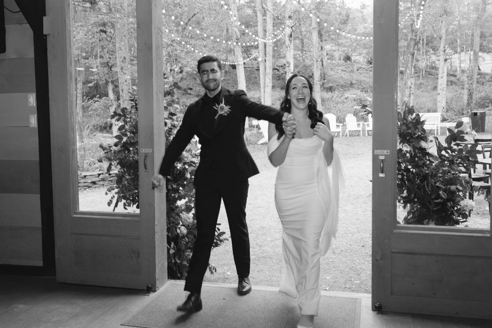 A couple in wedding attire entering a venue, holding hands and smiling, with string lights overhead and outdoor chairs visible in the background.
