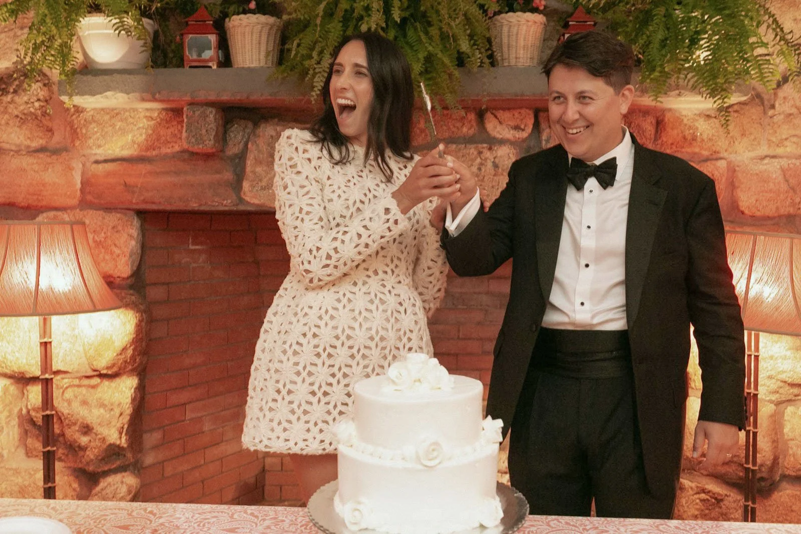 A couple dressed in formal attire at a celebration, with a woman in a white lace dress and a man in a black tuxedo, standing behind a white cake on a table, holding a knife together, smiling, in a warmly lit room with a brick and stone fireplace, pla