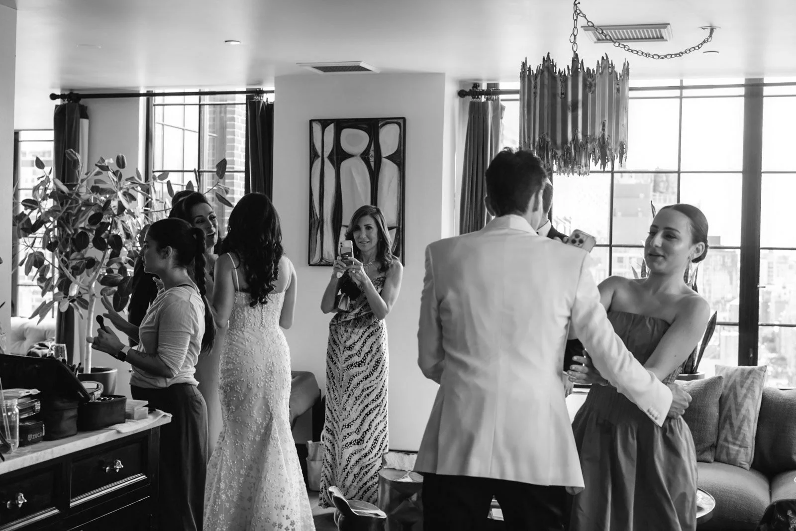 Group of people, mostly women, preparing and taking photos in a well-lit room with large windows, curtains, artwork, and modern decor.