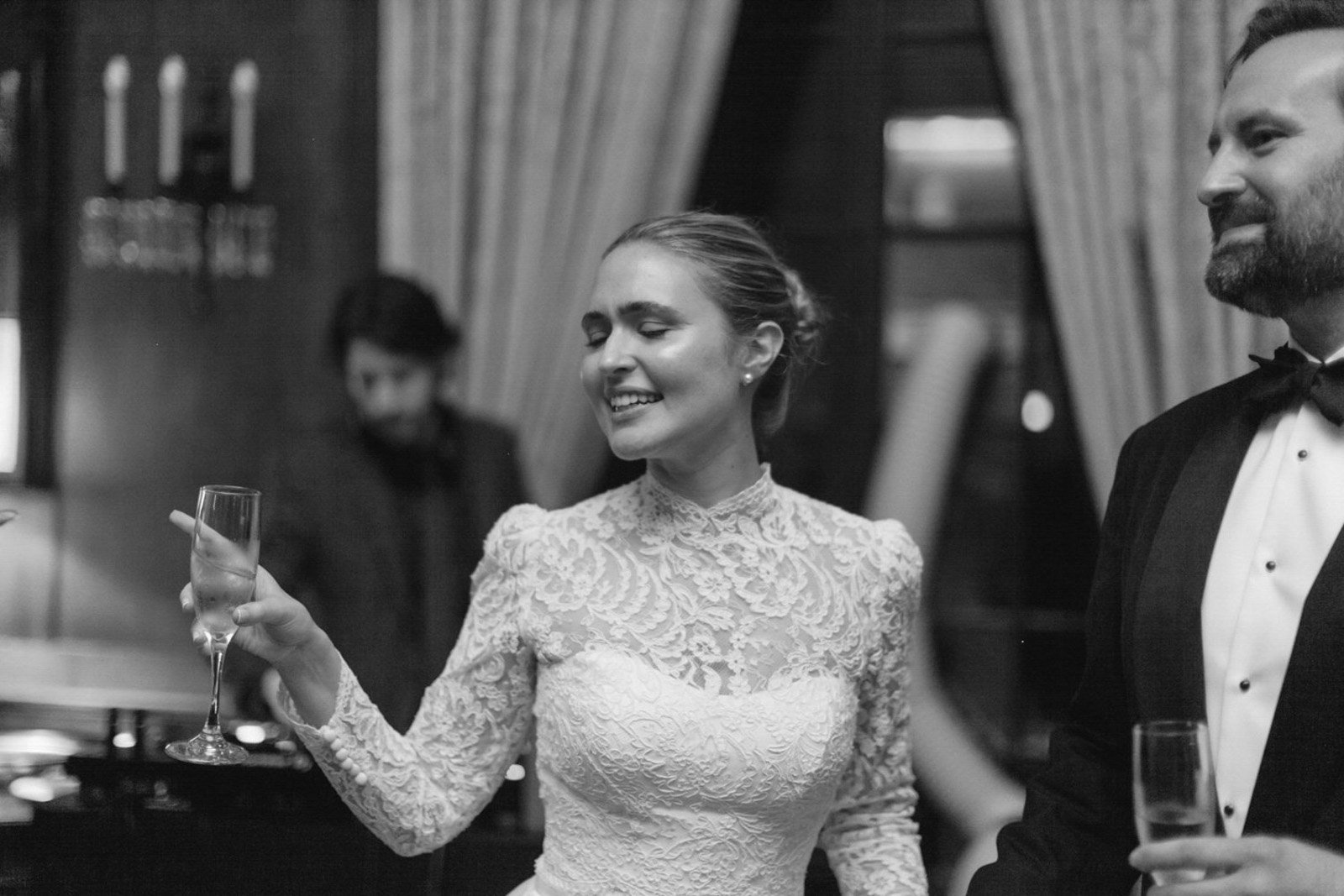 Black and white photo of a woman in a lace wedding dress holding a champagne flute, smiling with eyes closed, standing next to a man in a tuxedo at a formal event.