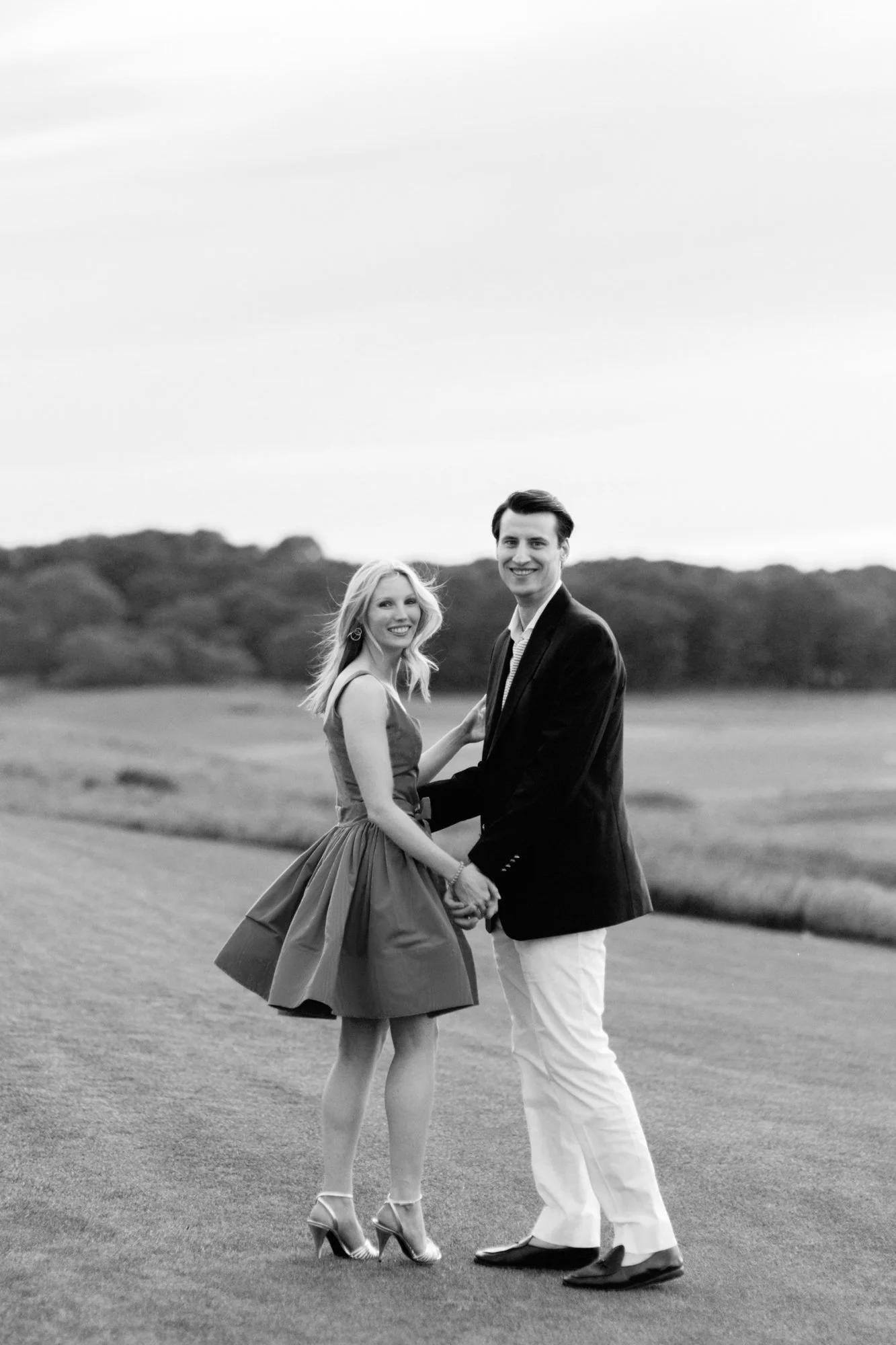 A black and white photo of a couple holding hands and smiling outdoors on a grassy field. The woman is wearing a dress and high heels, and the man is dressed in a jacket and pants.