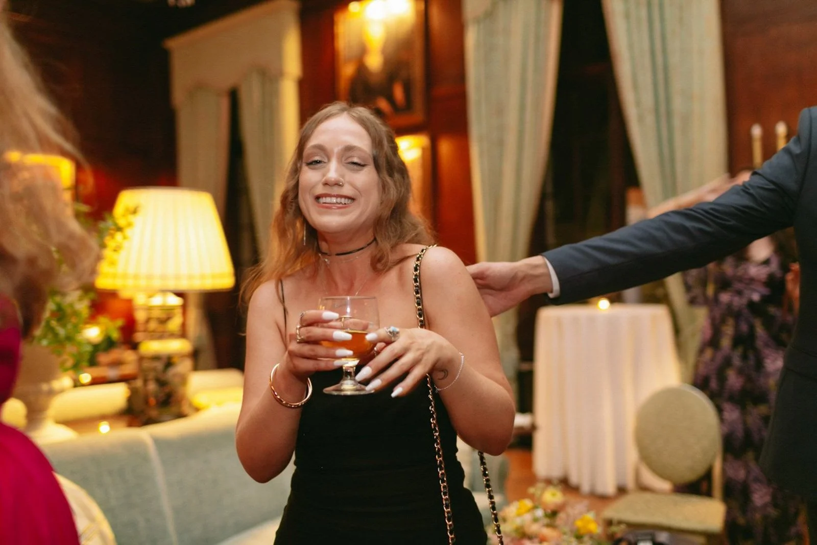 Woman with red hair smiling, holding a glass of amber-colored drink, in a warmly lit living room with lamps, curtains, and floral arrangements.
