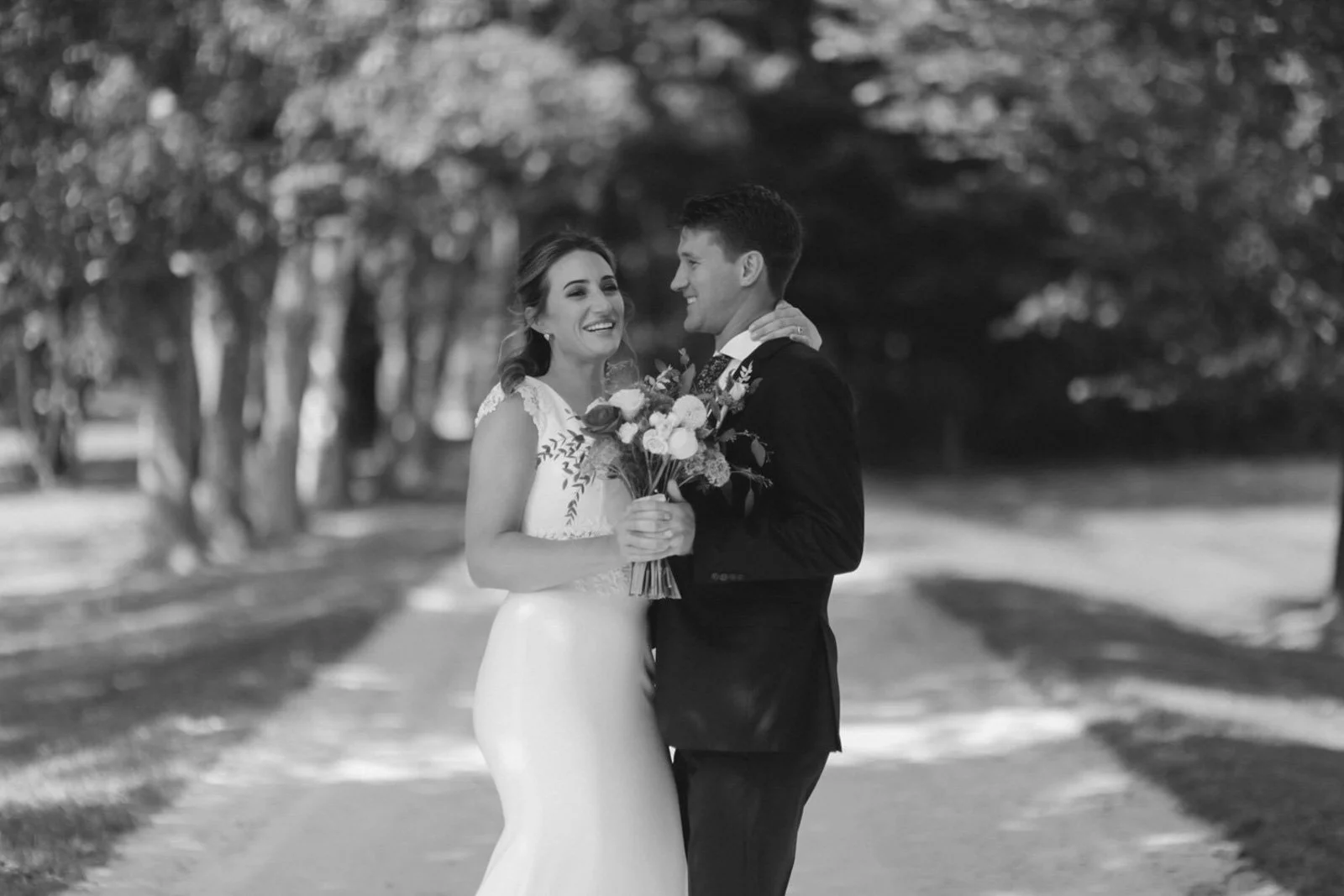 A black and white photo of a smiling bride and groom holding a bouquet of flowers, standing on a path in a park, embracing each other.