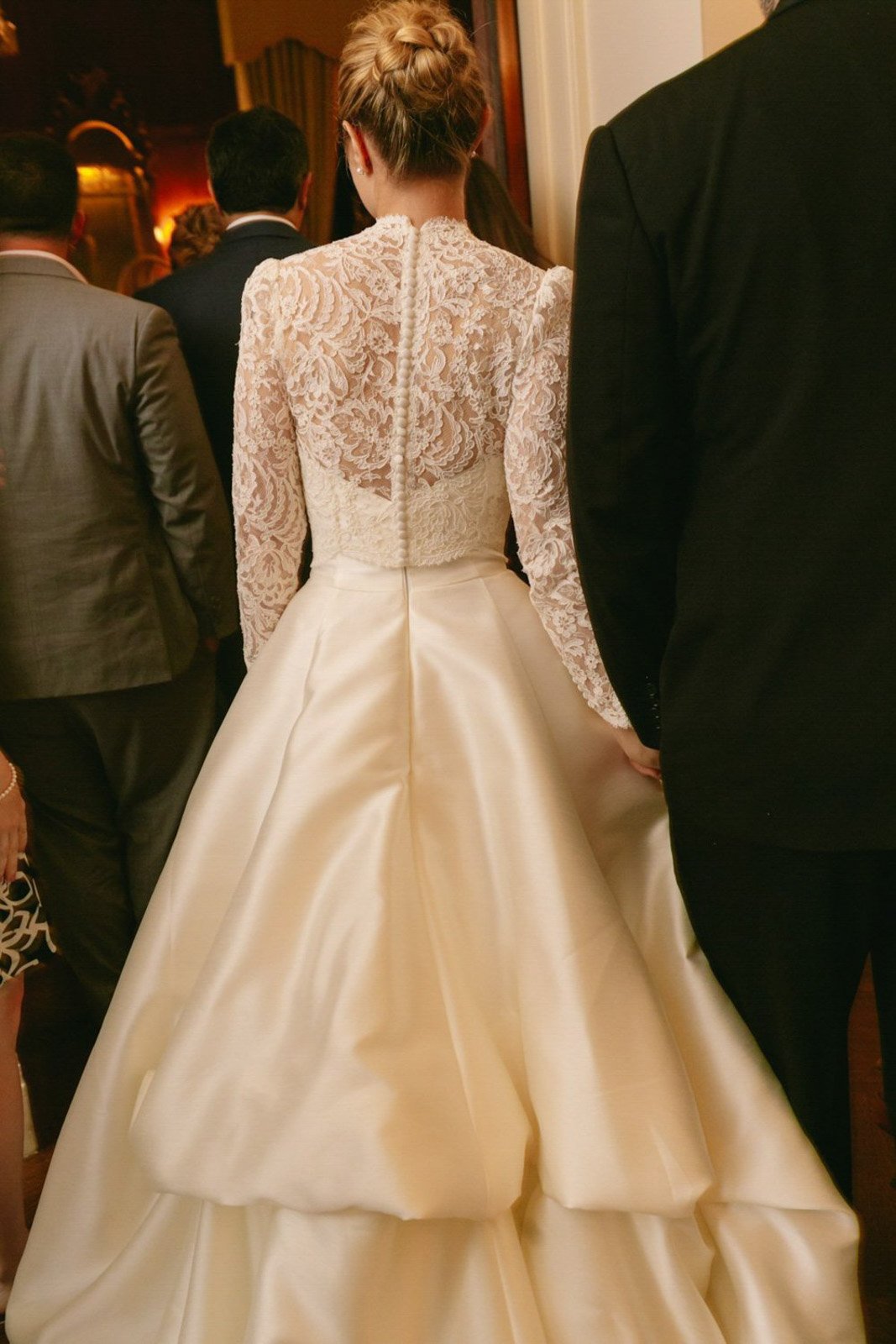 Back view of a bride in a white satin wedding gown with a lace top, surrounded by people in formal attire at an indoor wedding or formal event.