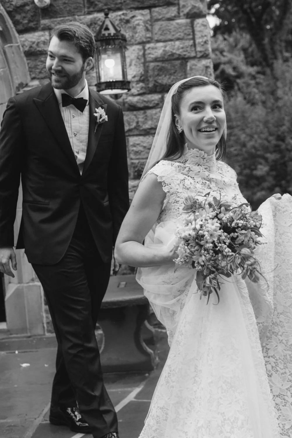 A bride and groom at a wedding, with the bride holding a bouquet of flowers and the groom wearing a tuxedo, outside stone building with lanterns.