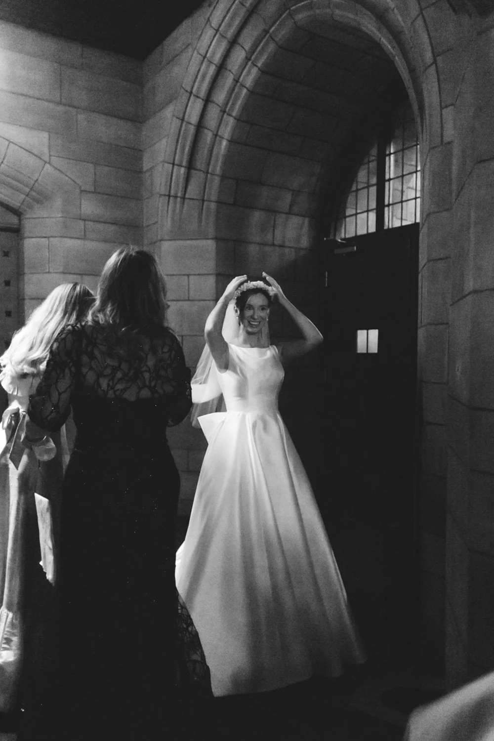 A bride in a white wedding dress and veil smiling and adjusting her headpiece inside a stone-walled building, with two women standing nearby.