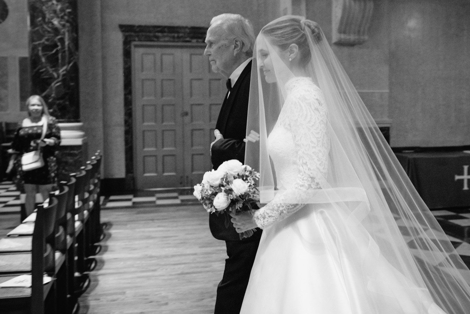 A bride walking down the aisle holding a bouquet, accompanied by an older man, in a church. The bride is wearing a lace wedding dress and veil. Seated guests are visible in the background.
