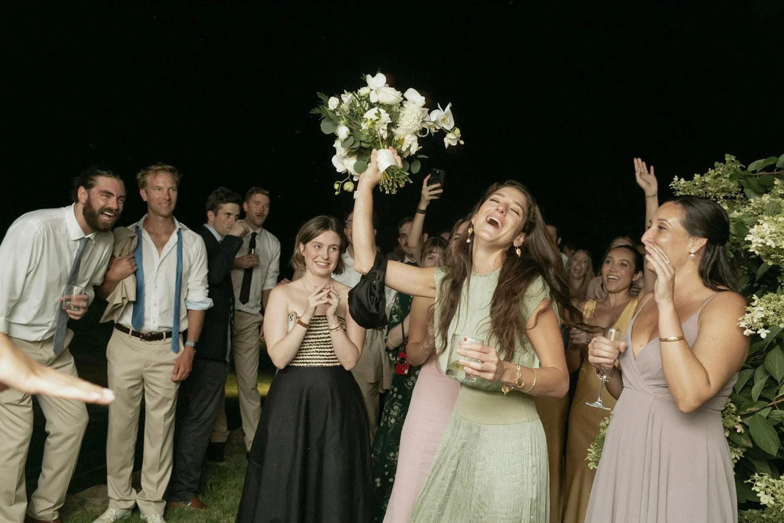 A group of people celebrating outdoors at night, with a woman holding a bouquet of white flowers raised in the air, smiling and laughing, surrounded by friends dressed in semi-formal attire.