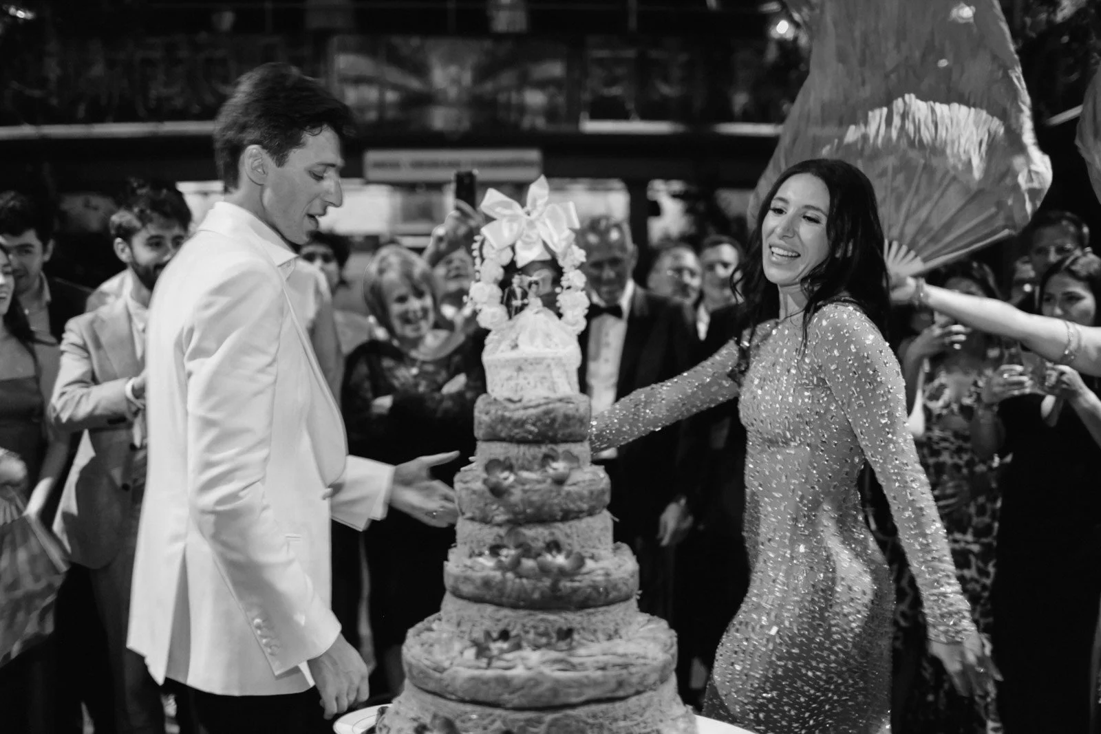 A woman and a man in formal attire cutting a wedding cake at a celebration with guests around them.
