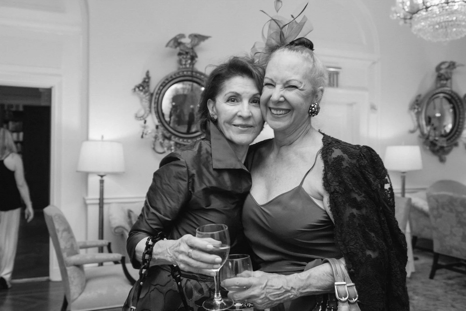 Two women smiling and holding wine glasses at an indoor social gathering, with elegant decor and mirrors in the background.