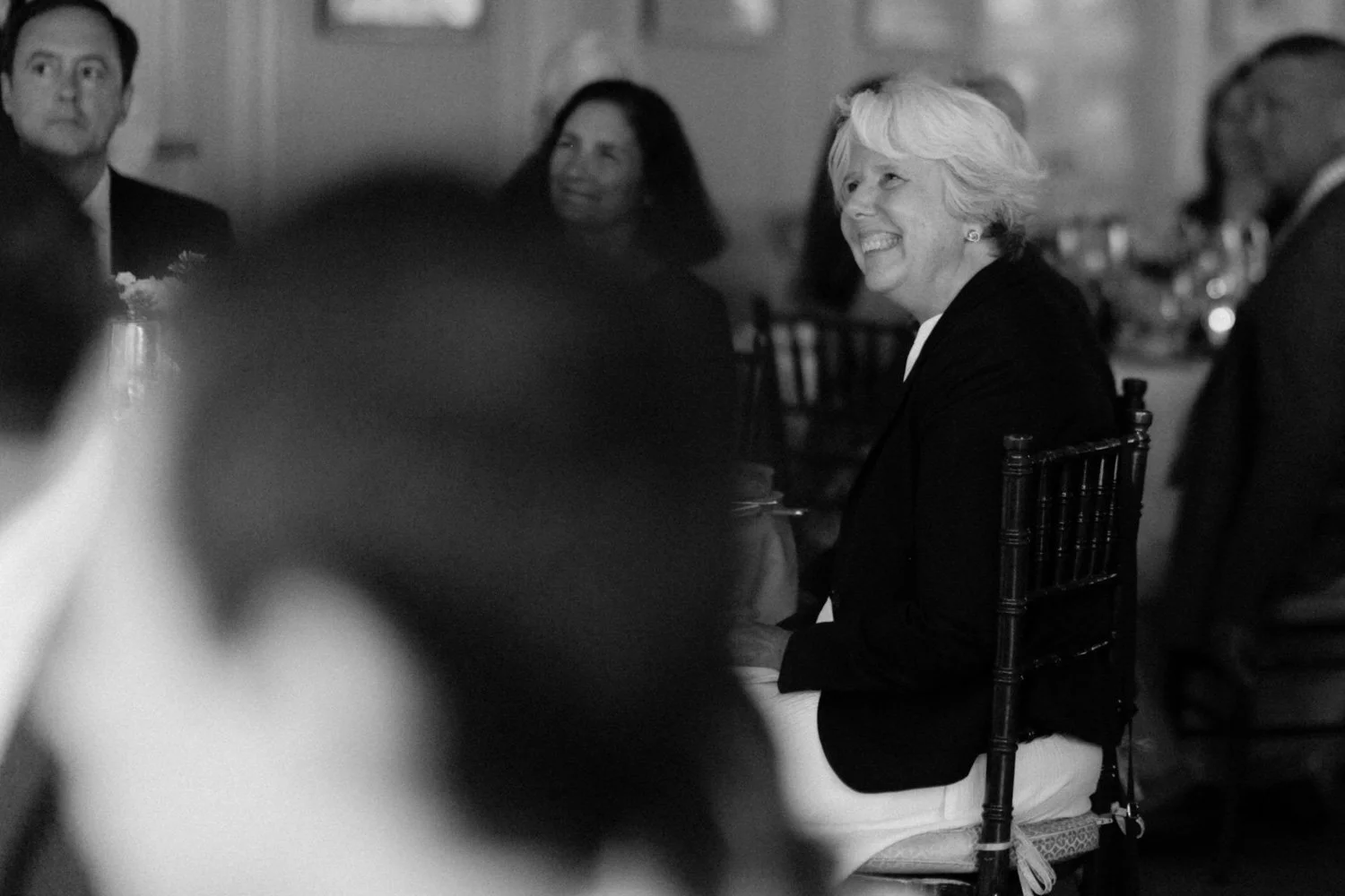 A group of people seated at a formal event, with a focus on a woman smiling and looking to her right. The image is in black and white.
