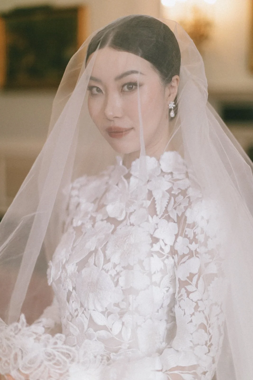 A woman in a white wedding dress with floral lace details, wearing a veil and elegant earrings, smiling softly.