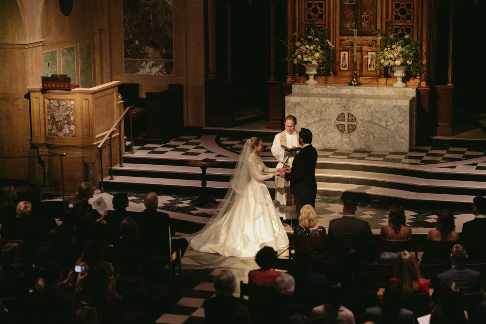 A wedding ceremony inside a church, with a bride and groom holding hands and exchanging vows, officiated by a priest standing at the altar. The altar is decorated with flowers and religious icons, and guests are seated and watching.