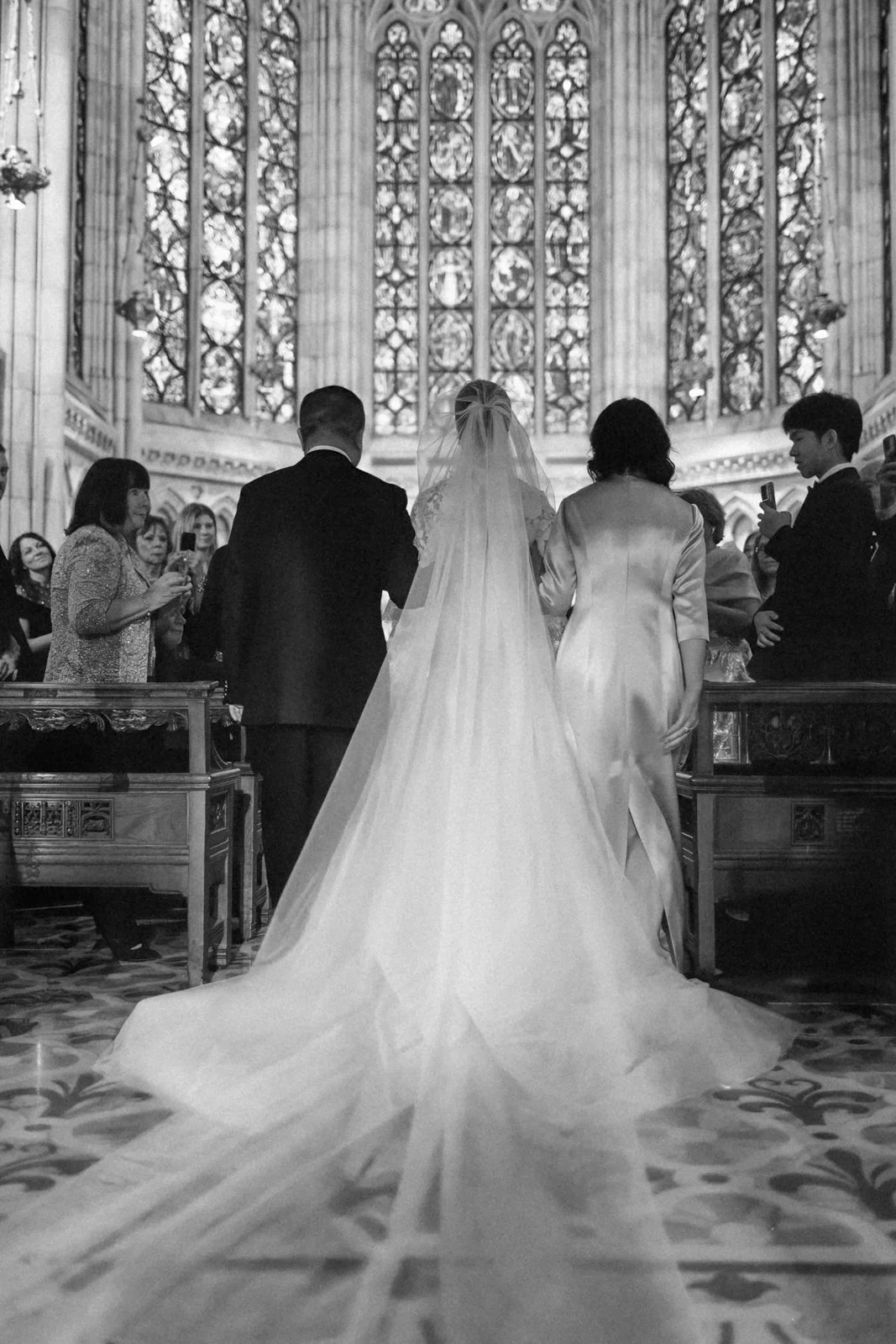A black and white photo of a bride and groom holding hands at the altar during their wedding ceremony inside a church with stained glass windows. Guests are taking photos and watching the couple.
