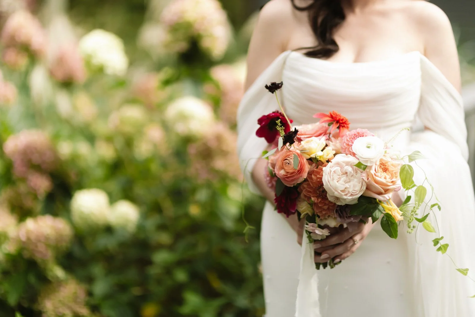 A woman in a white wedding dress holding a colorful bouquet of flowers outdoors, with blurred pinks and greens in the background.