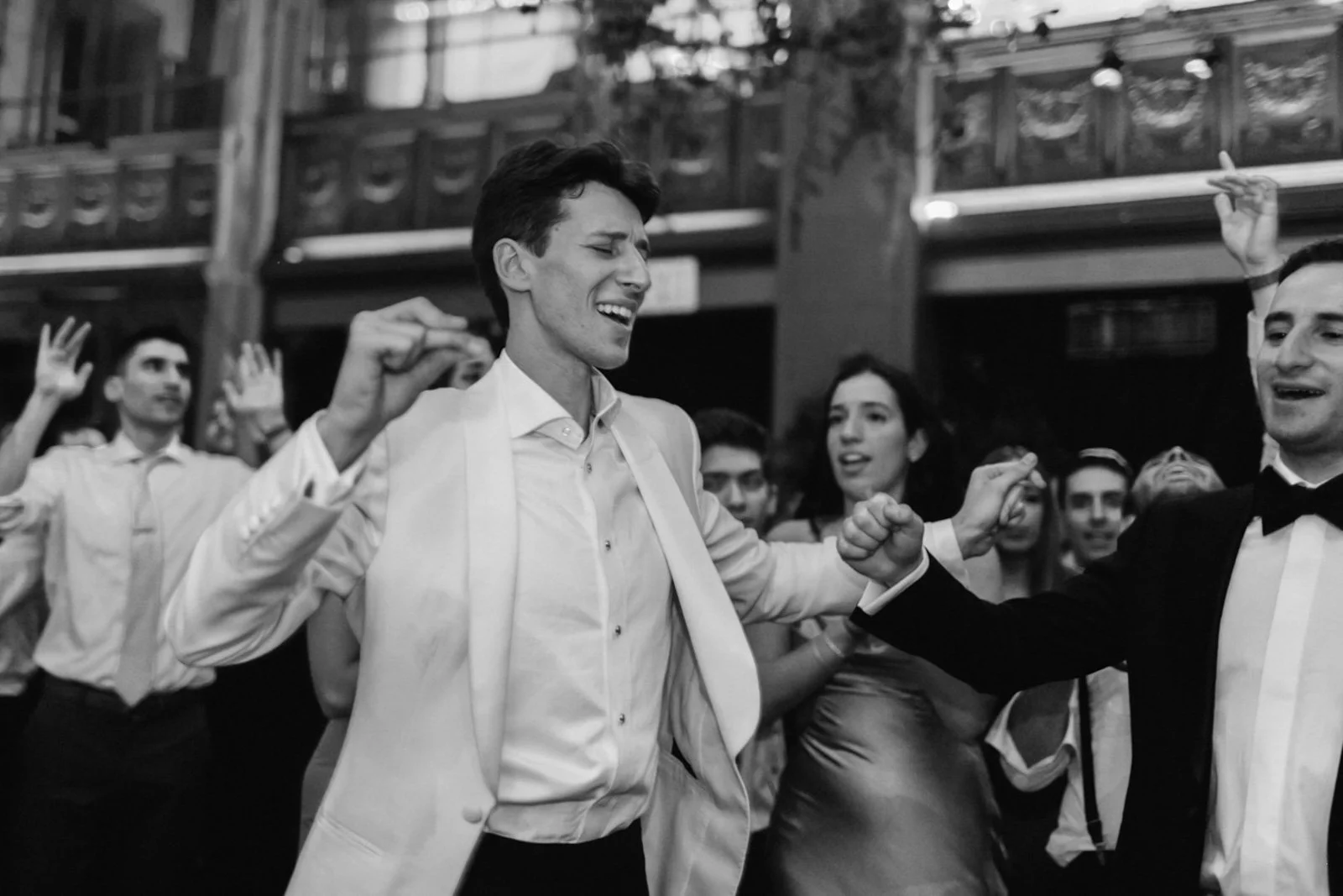 A black and white photo of people dancing at a party or celebration, with two men in the foreground engaging in a playful arm wrestling or fist bump, smiling and enjoying themselves. Other people are visible in the background, some raising their hand