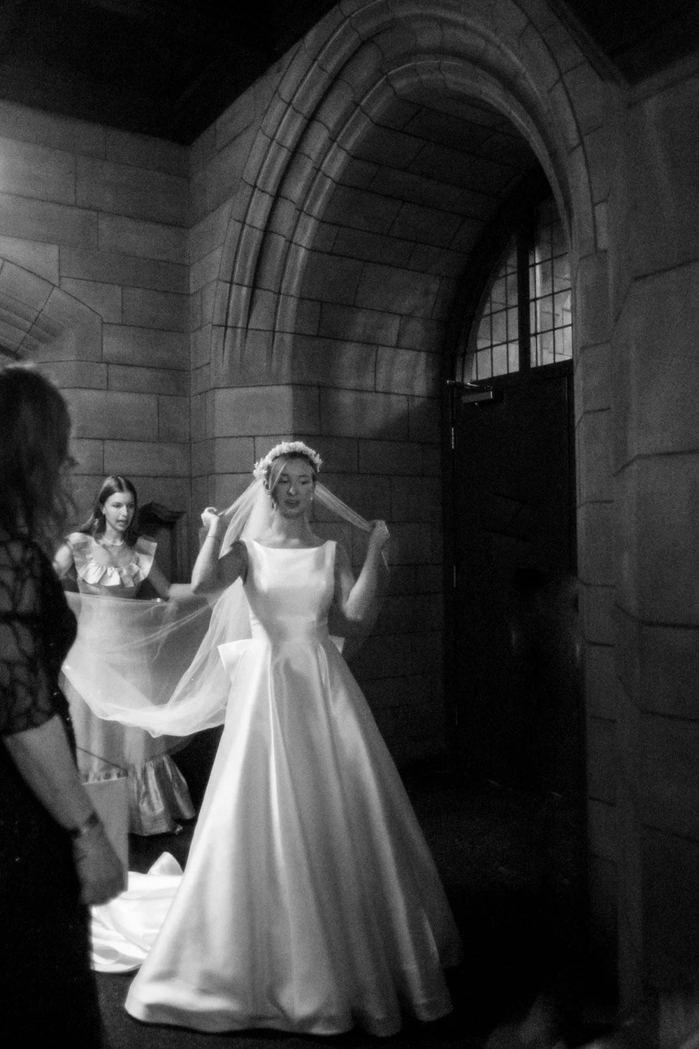 A bride in a wedding dress is adjusting her veil inside a stone church while two women assist her.