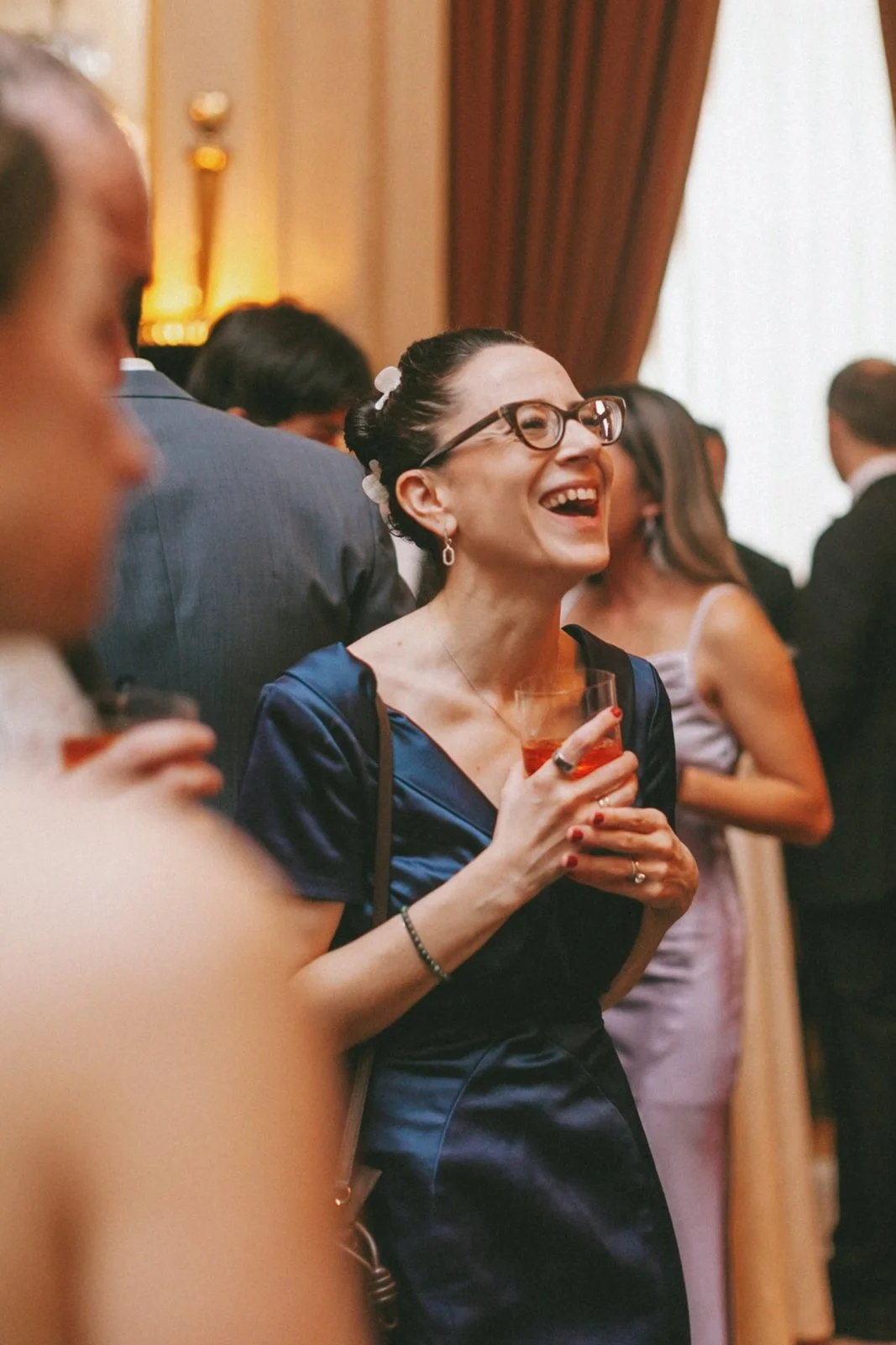 A woman with glasses and dark hair styled in an updo, smiling and holding a glass of red drink at a social gathering, surrounded by other people in formal attire.