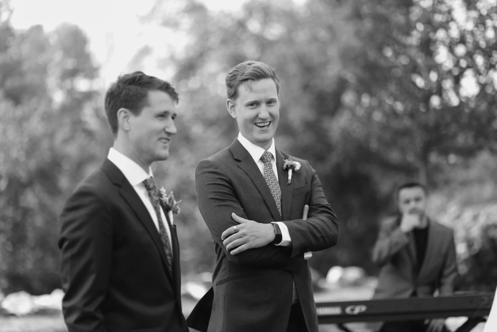 Two men in suits and a third person in the background standing outdoors during a formal event, likely a wedding, with trees in the background.