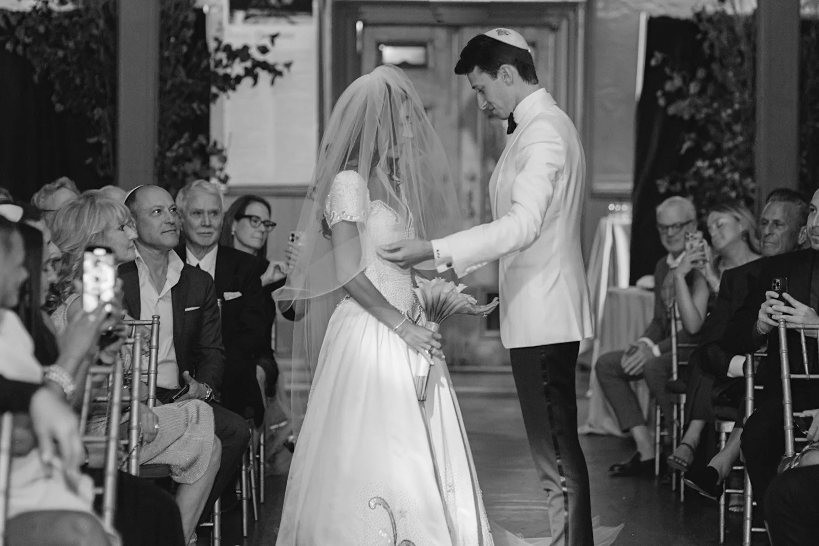 A black-and-white photo of a wedding ceremony with a bride and groom standing close, the bride holding a bouquet, in an ornate room with seated guests watching as the groom places a ring on the bride's finger.