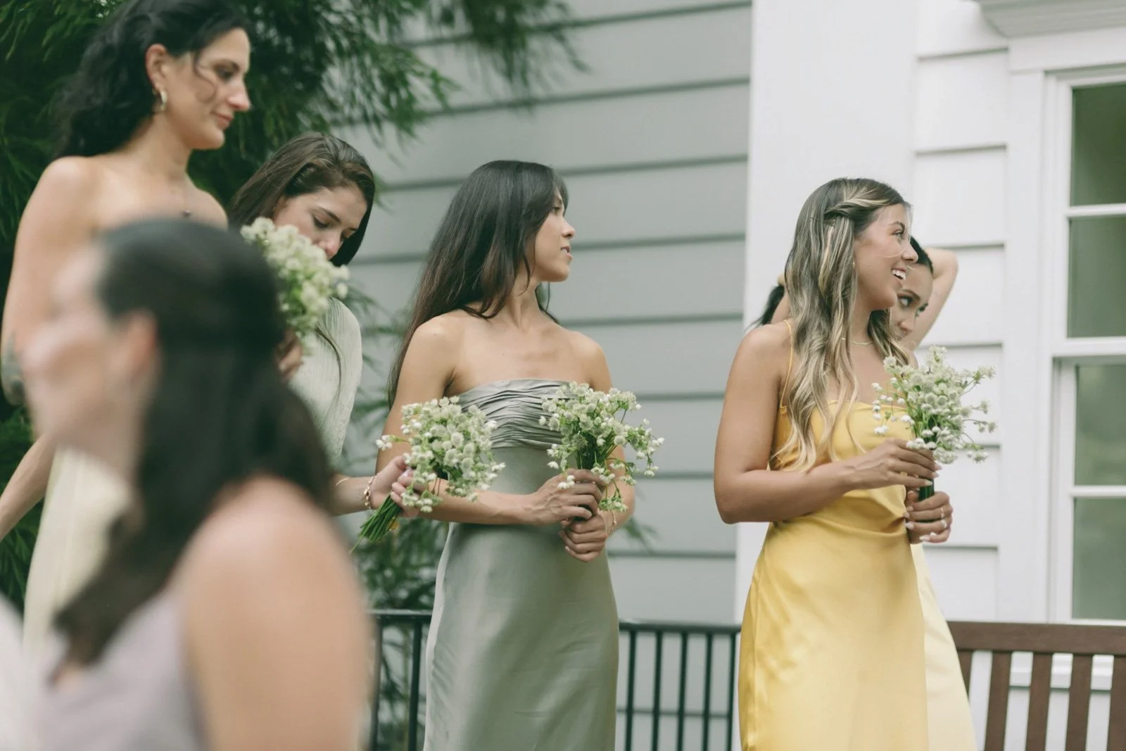 A group of women in elegant dresses holding bouquets, attending a wedding or similar event outdoors, near a white building with large windows.