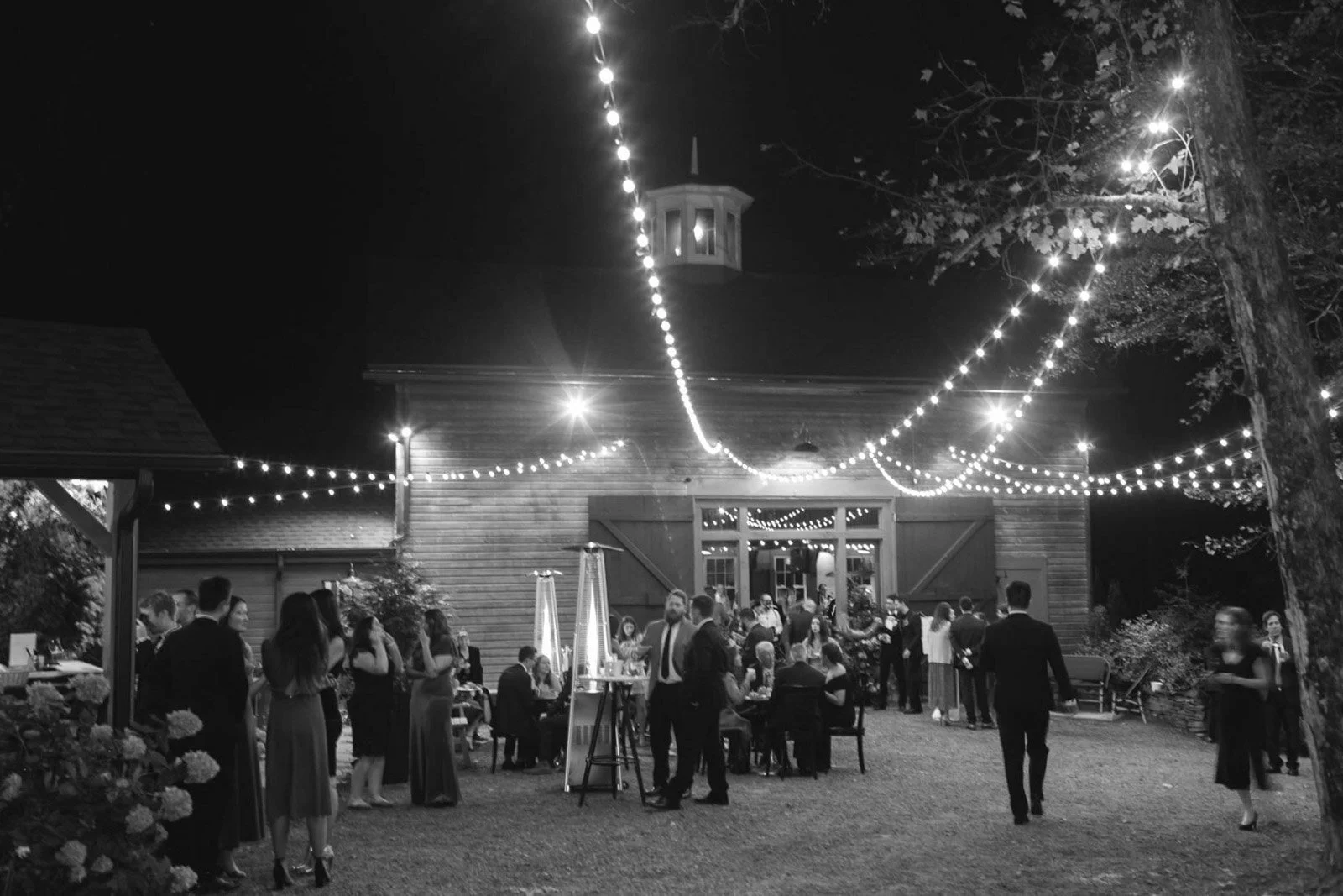 Black and white photo of an outdoor nighttime party with people dressed in formal attire, string lights hanging above, and a building with open doors in the background.