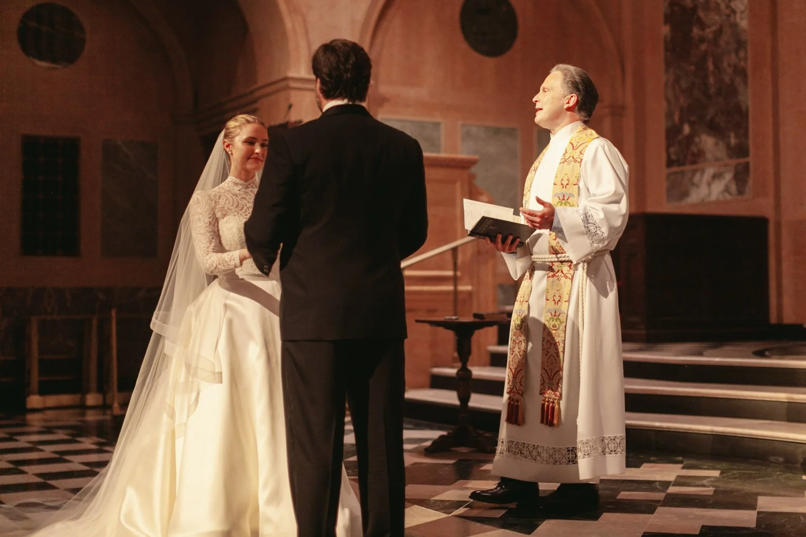A wedding ceremony taking place in a church with a bride in a lace wedding dress and long veil, a groom in a black tuxedo, and a priest officiating.