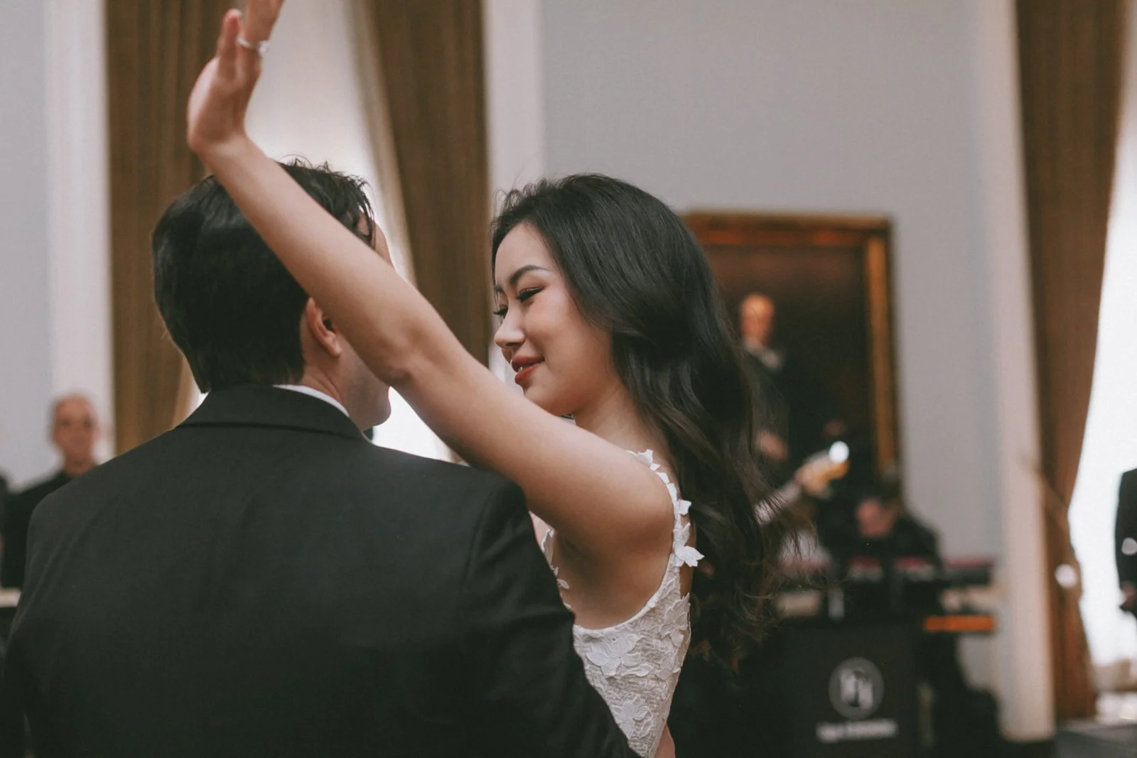 A woman and a man dancing closely at a formal event, with the woman's arm raised, smiling, in an elegant room with curtains and a band in the background.