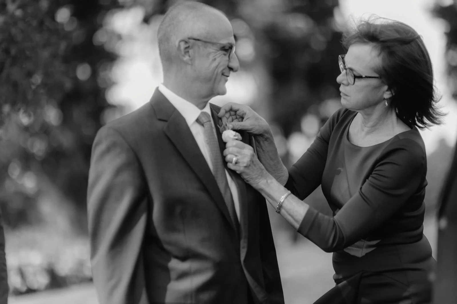 A woman pins a boutonniere on a smiling man's suit jacket during an outdoor event.