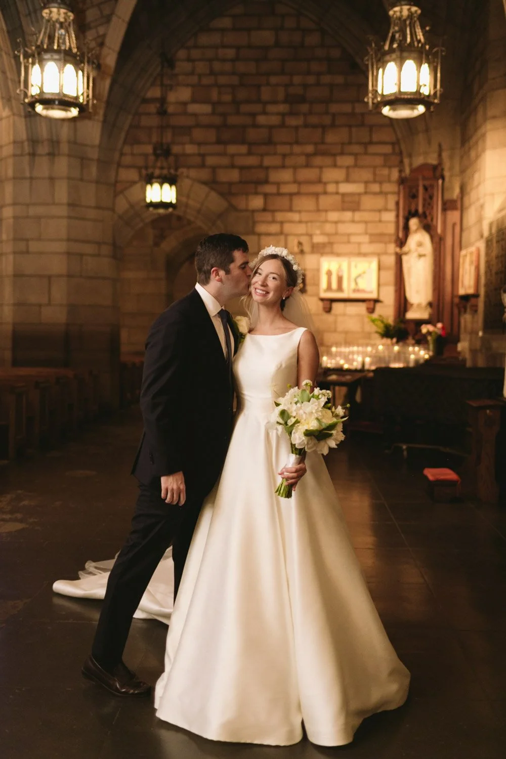 A bride and groom in a church, with the groom kissing the bride on the temple. The bride is smiling and holding a bouquet of white flowers, wearing a white wedding gown and a floral headband. The church has stone walls, hanging lanterns, and religiou