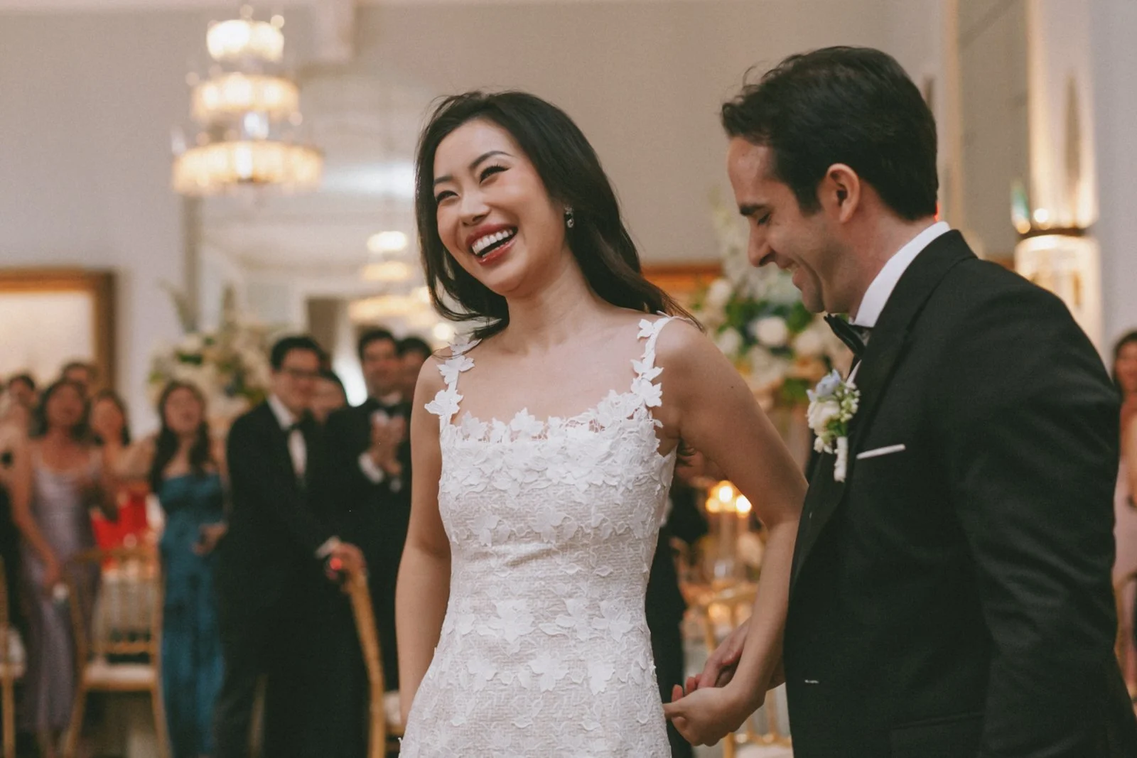 A bride and groom holding hands at their wedding ceremony, surrounded by guests in a decorated indoor venue.