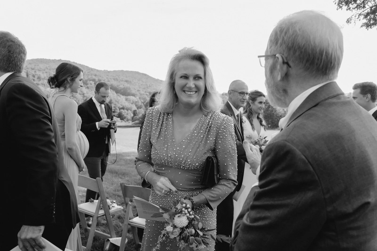 A group of people at an outdoor event, with a woman smiling and holding a bouquet of flowers in the center, surrounded by others engaged in conversation near a body of water with hills in the background.